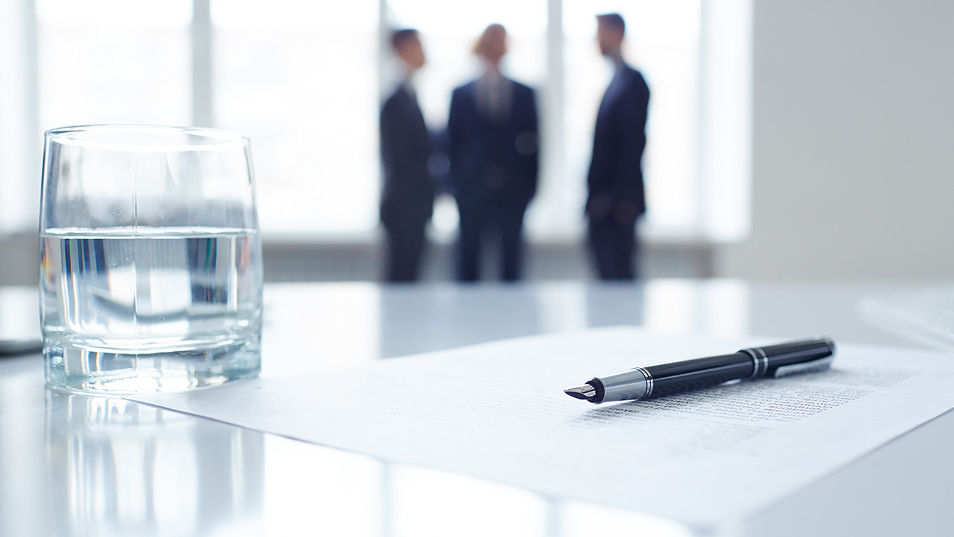 Image of business document, pen and glass of water at workplace with group of colleagues on background